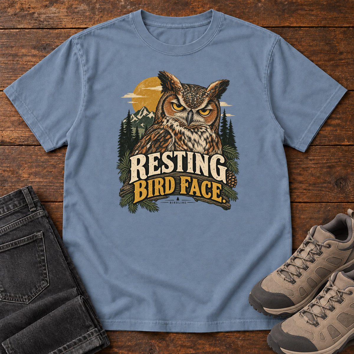 Blue Jean Resting Bird Face T-Shirt with a yellow-eyed, tufted owl with pine branches, mountains and a golden sun; front flat lay on wood.