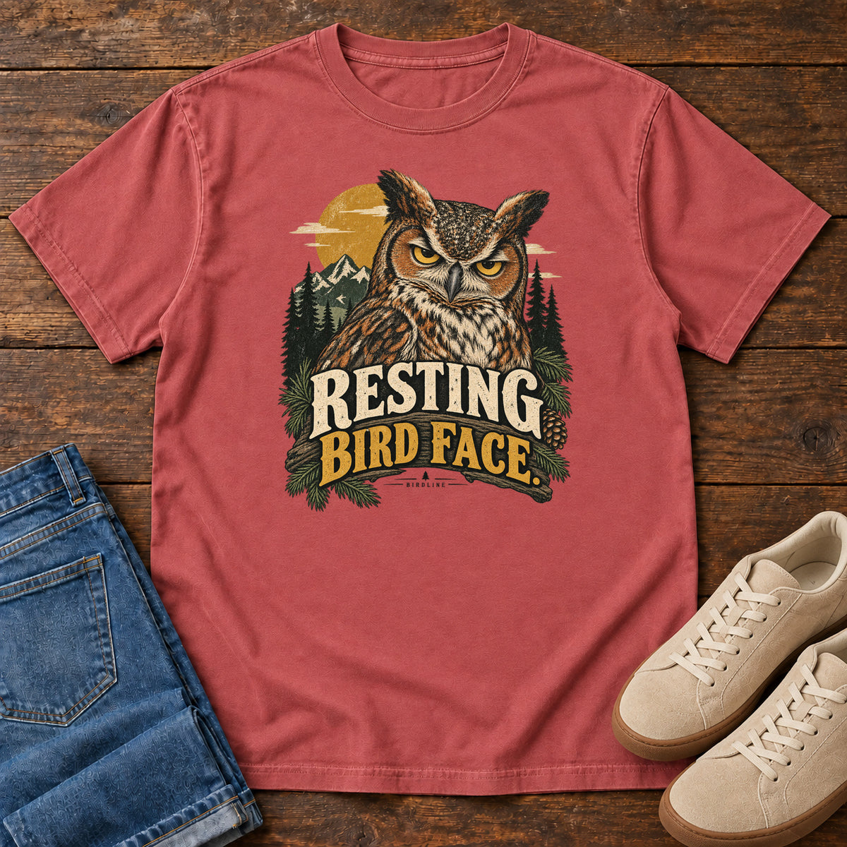 Crimson Resting Bird Face T-Shirt with a yellow-eyed, tufted owl with pine branches, mountains and a golden sun; front flat lay on wood.
