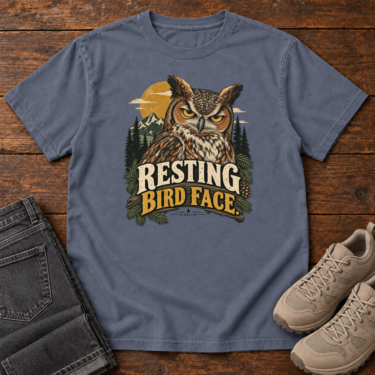 Denim Resting Bird Face T-Shirt with a yellow-eyed, tufted owl with pine branches, mountains and a golden sun; front flat lay on wood.