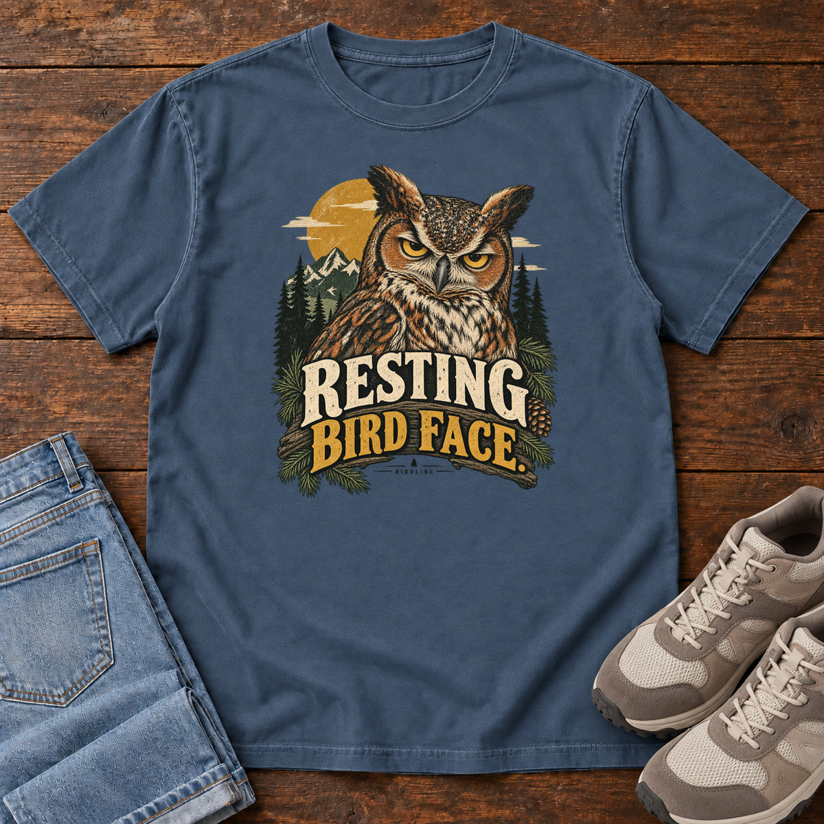 Midnight Resting Bird Face T-Shirt with a yellow-eyed, tufted owl with pine branches, mountains and a golden sun; front flat lay on wood.