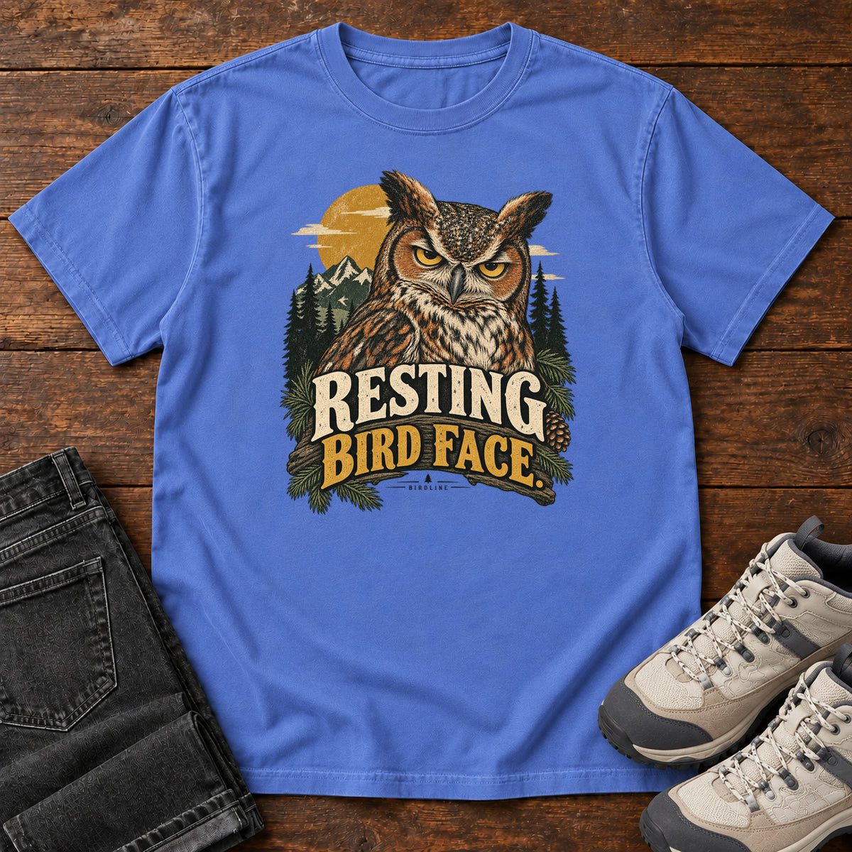 Mystic Blue Resting Bird Face T-Shirt with a yellow-eyed, tufted owl with pine branches, mountains and a golden sun; front flat lay on wood.