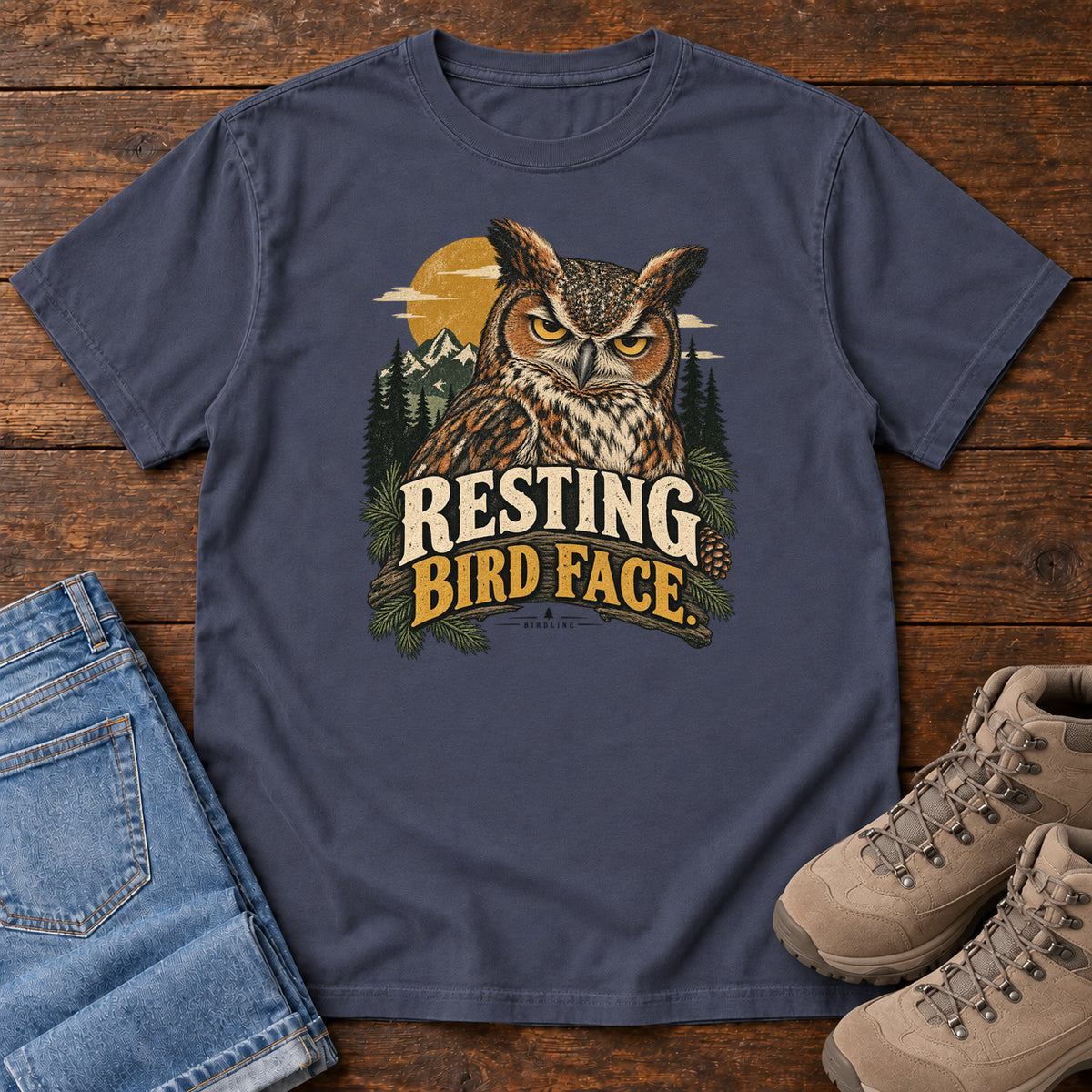 Navy Resting Bird Face T-Shirt with a yellow-eyed, tufted owl with pine branches, mountains and a golden sun; front flat lay on wood.