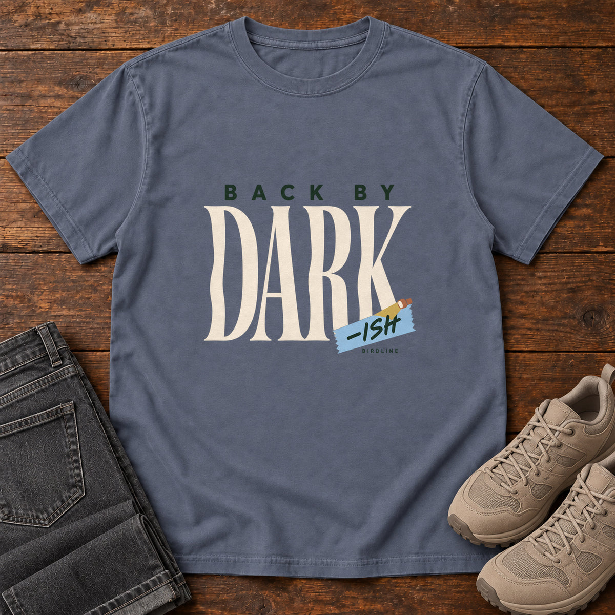 Denim Back by Dark-ish T-Shirt with tall cream Dark lettering and a small blue -ish label, front flat lay on wood.