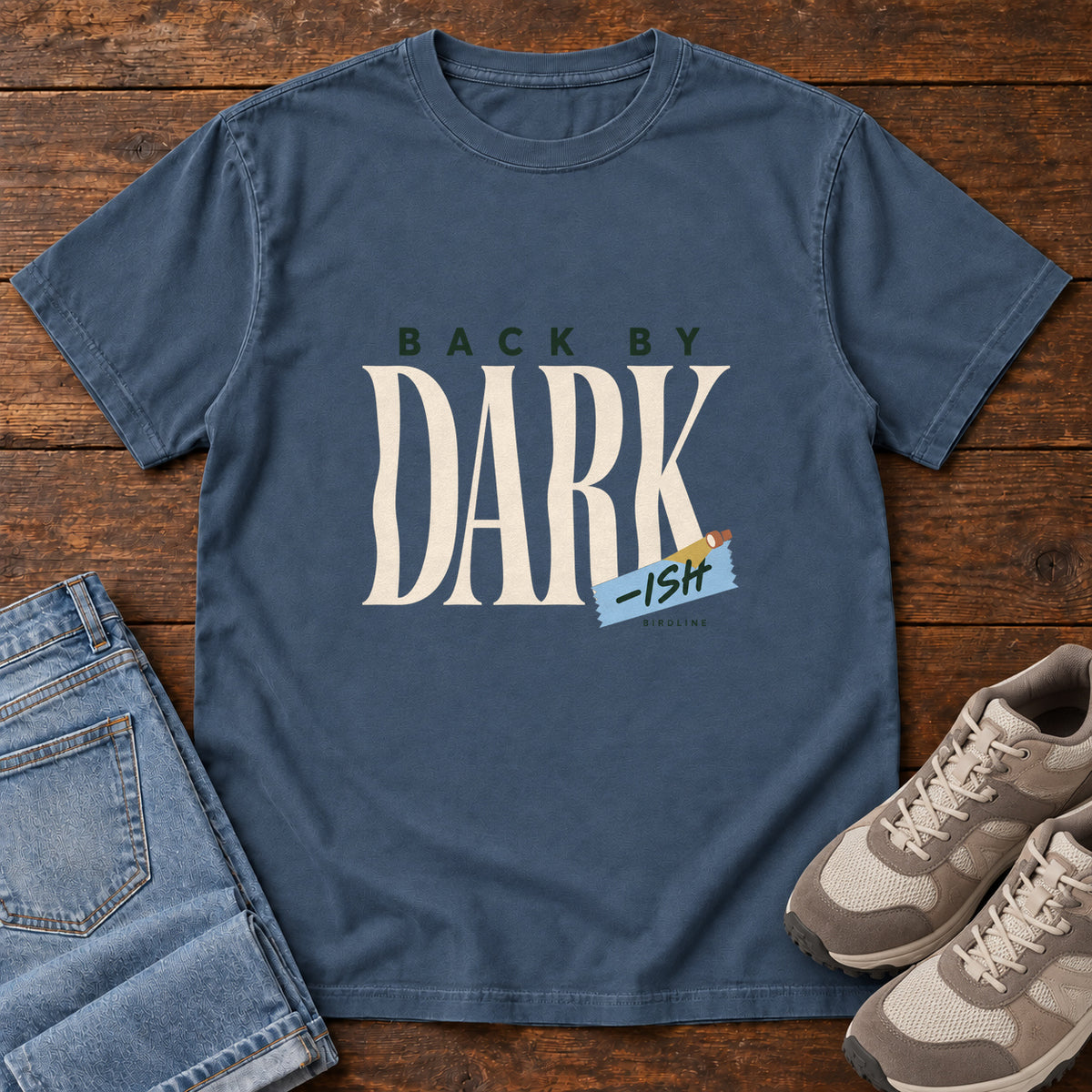 Midnight Back by Dark-ish T-Shirt with tall cream Dark lettering and a small blue -ish label, front flat lay on wood.