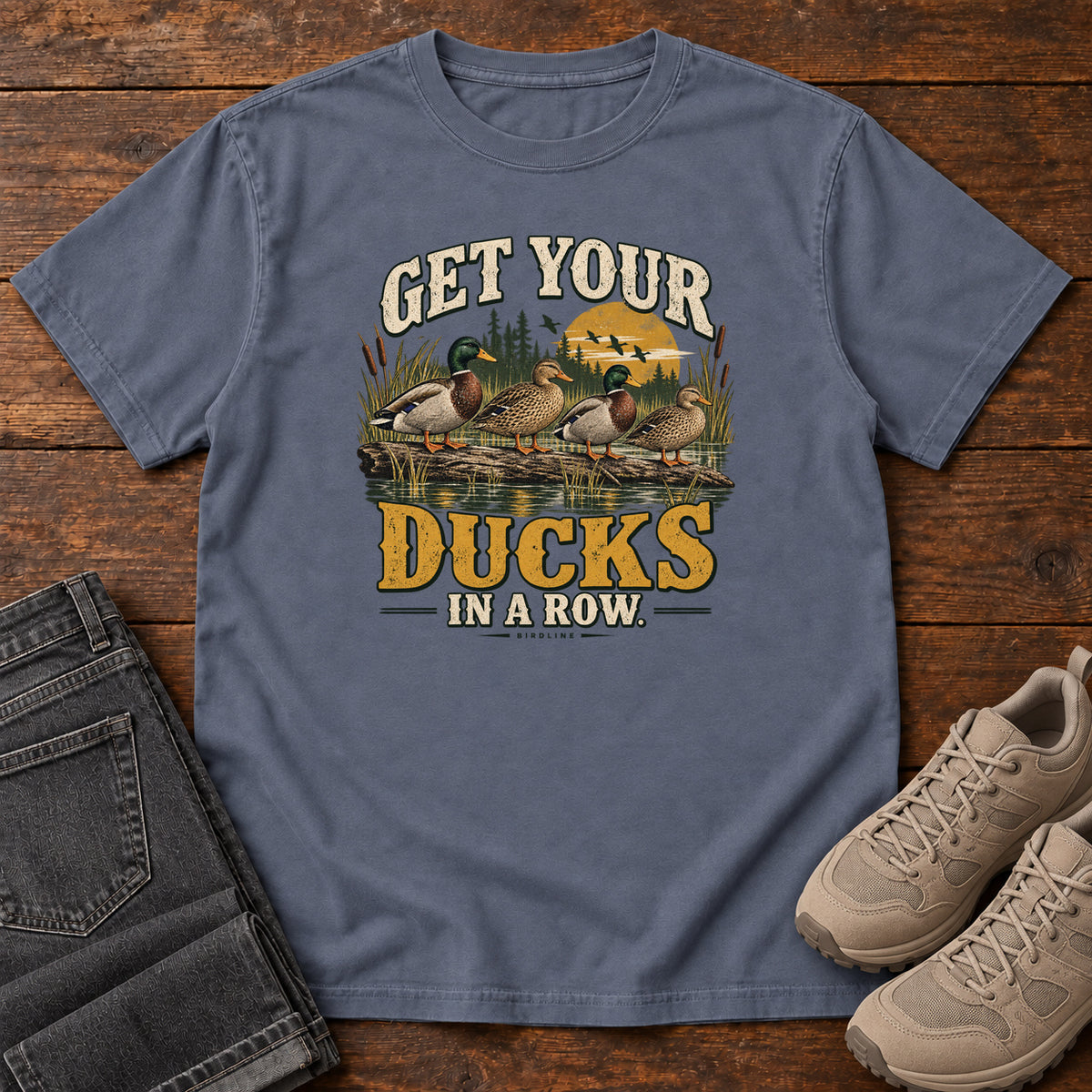 Get Your Ducks in a Row T-Shirt in Denim, front laid flat on wood, featuring a row of ducks among reeds and water beneath cream-and-gold slogan lettering.