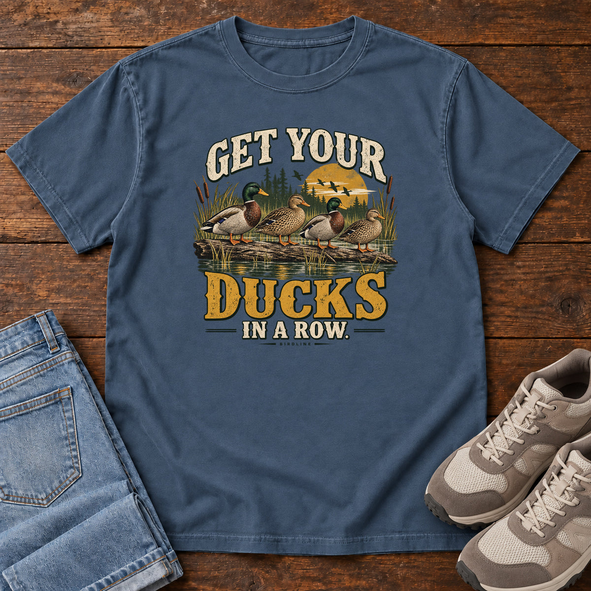 Get Your Ducks in a Row T-Shirt in Midnight, front laid flat on wood, featuring a row of ducks among reeds and water beneath cream-and-gold slogan lettering.