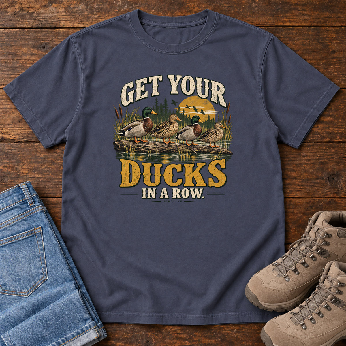 Get Your Ducks in a Row T-Shirt in Navy, front laid flat on wood, featuring a row of ducks among reeds and water beneath cream-and-gold slogan lettering.