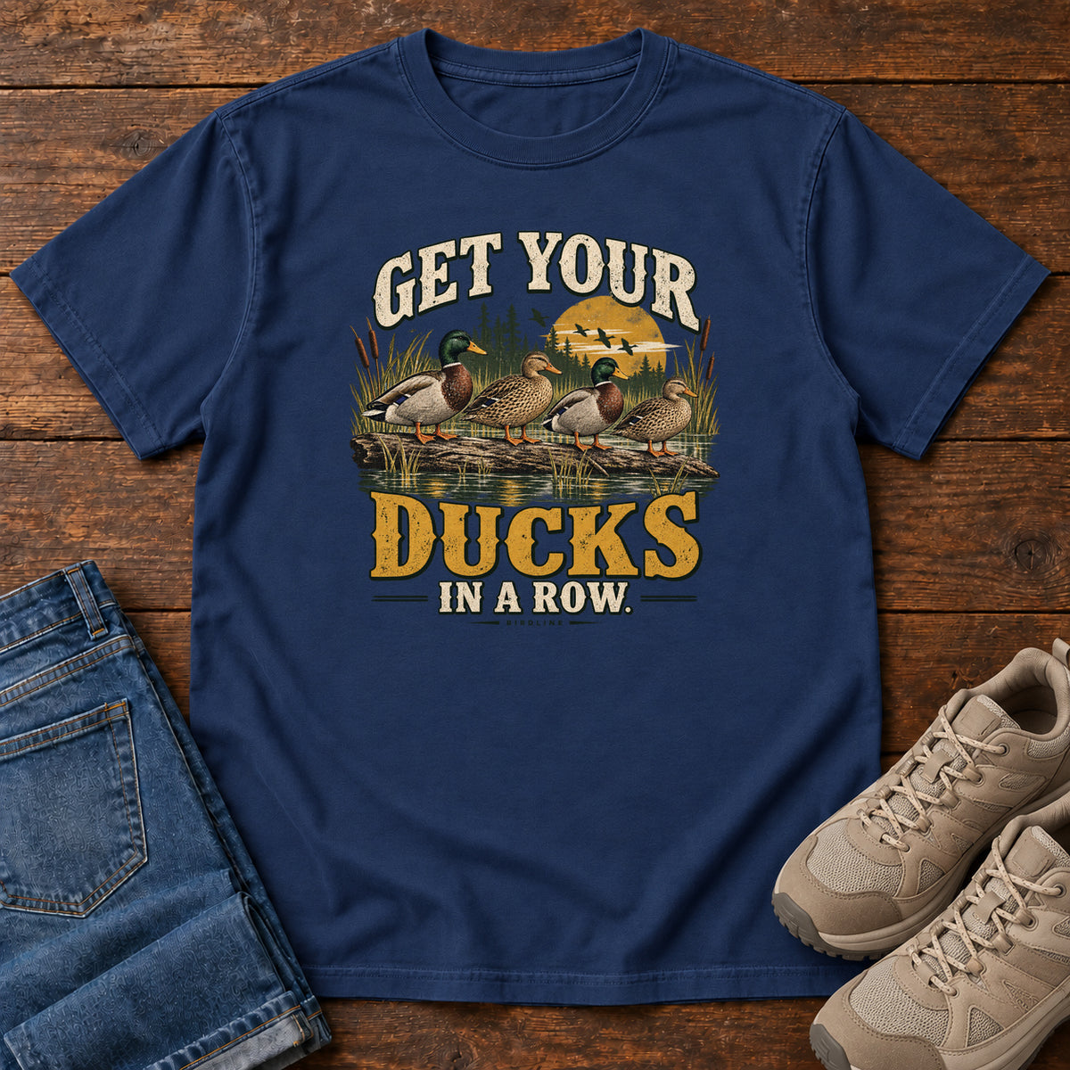 Get Your Ducks in a Row T-Shirt in True Navy, front laid flat on wood, featuring a row of ducks among reeds and water beneath cream-and-gold slogan lettering.