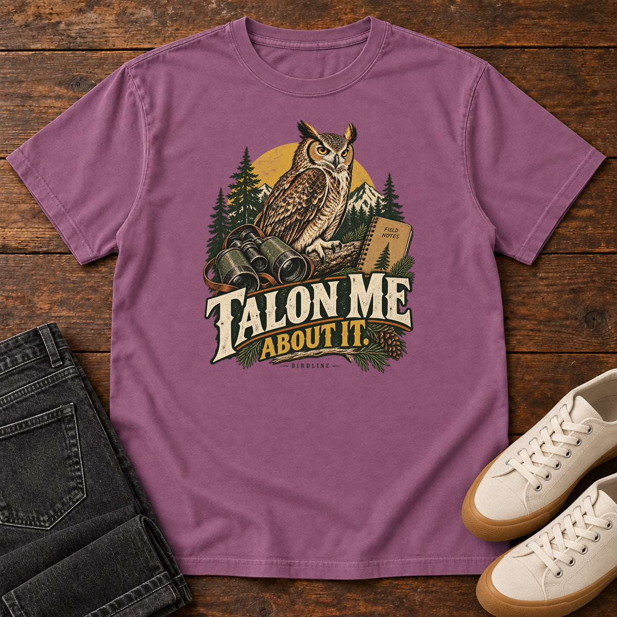 Berry Talon Me About It T-Shirt with an owl, binoculars and a field notebook against a forest backdrop, front flat lay on wood.