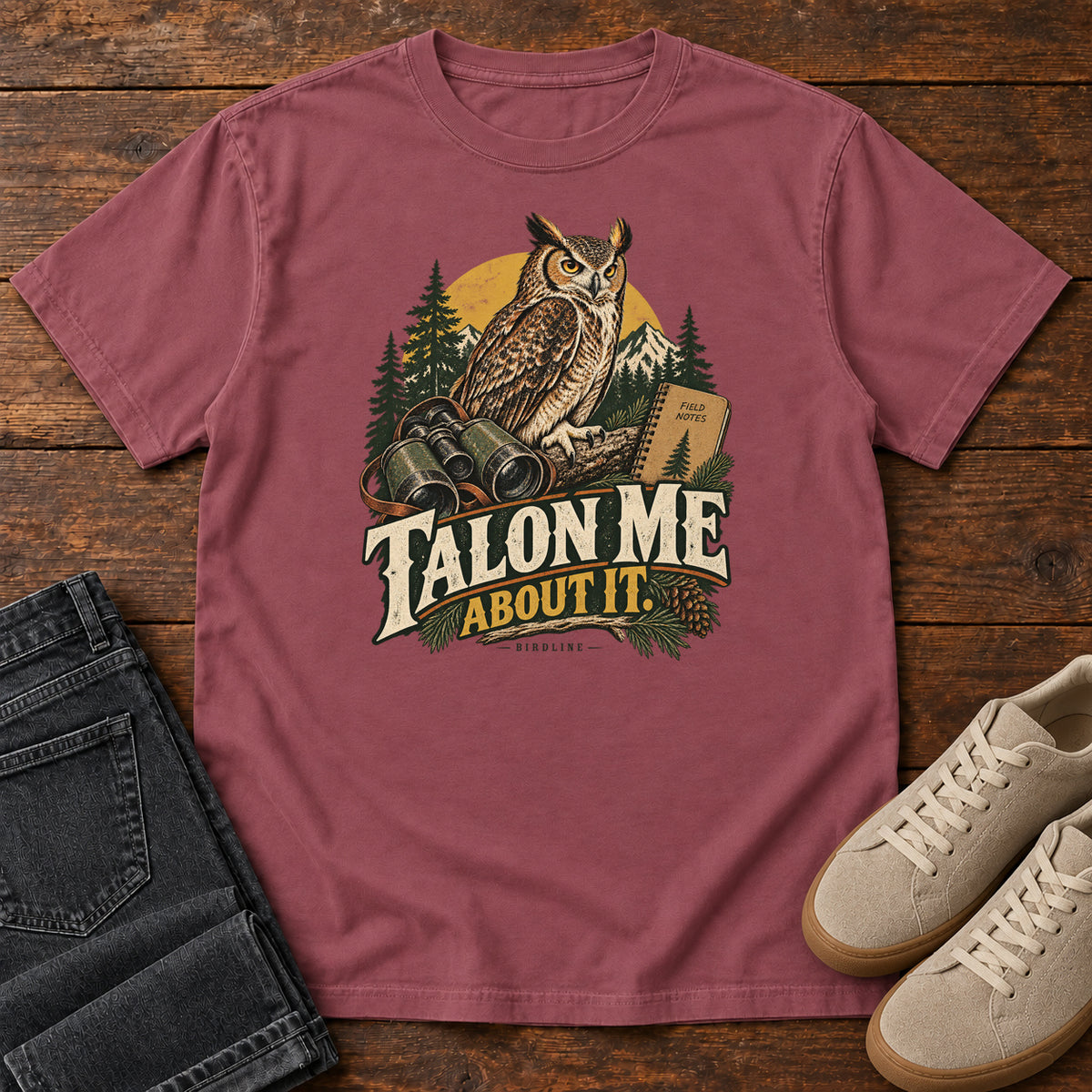 Brick Talon Me About It T-Shirt with an owl, binoculars and a field notebook against a forest backdrop, front flat lay on wood.