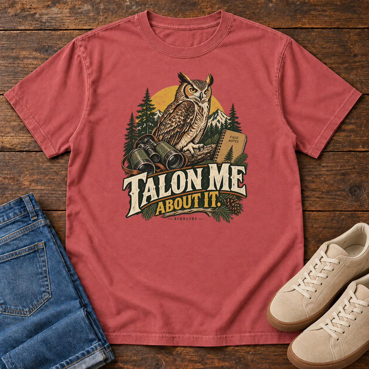 Crimson Talon Me About It T-Shirt with an owl, binoculars and a field notebook against a forest backdrop, front flat lay on wood.