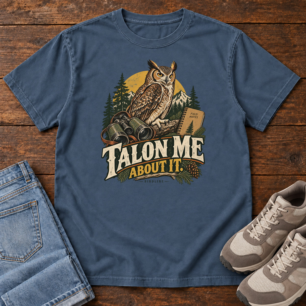 Midnight Talon Me About It T-Shirt with an owl, binoculars and a field notebook against a forest backdrop, front flat lay on wood.