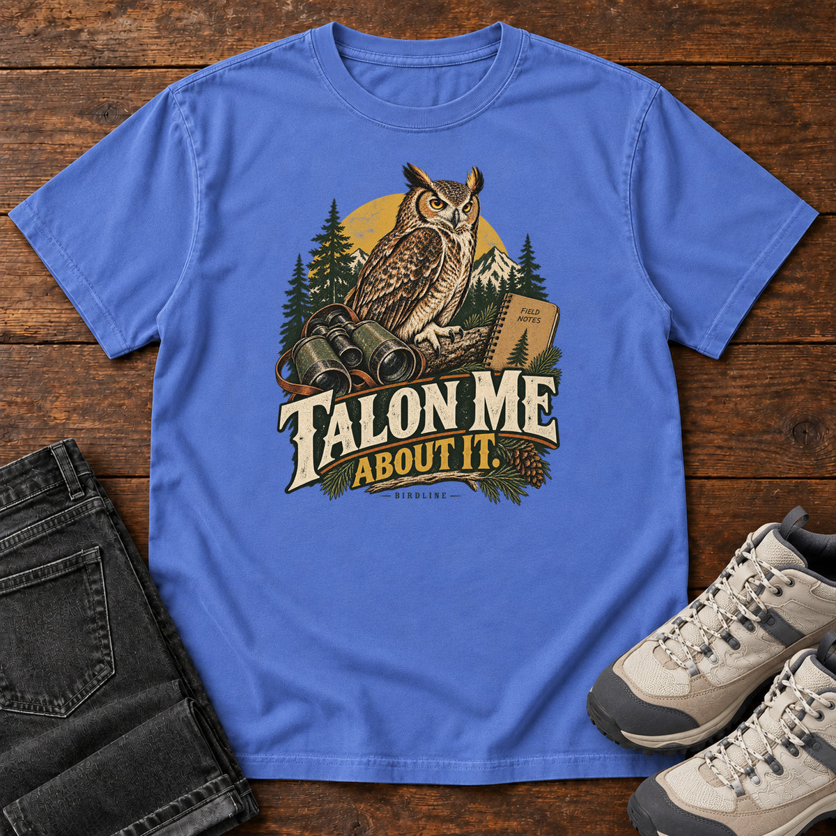 Mystic Blue Talon Me About It T-Shirt with an owl, binoculars and a field notebook against a forest backdrop, front flat lay on wood.