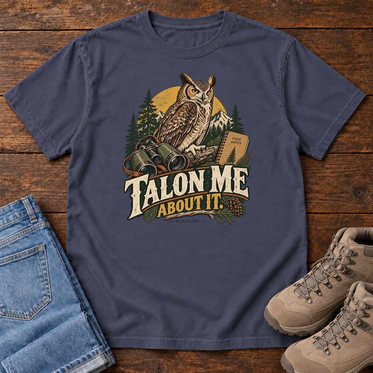 Navy Talon Me About It T-Shirt with an owl, binoculars and a field notebook against a forest backdrop, front flat lay on wood.