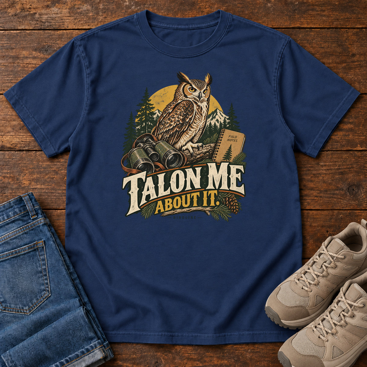 True Navy Talon Me About It T-Shirt with an owl, binoculars and a field notebook against a forest backdrop, front flat lay on wood.