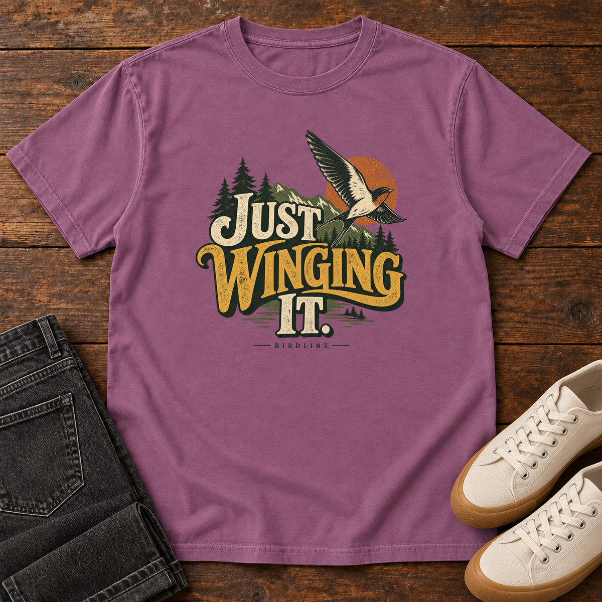 Berry Just Winging It T-Shirt with a flying bird, evergreens, an orange sun and cream and gold lettering; front flat lay on wood.