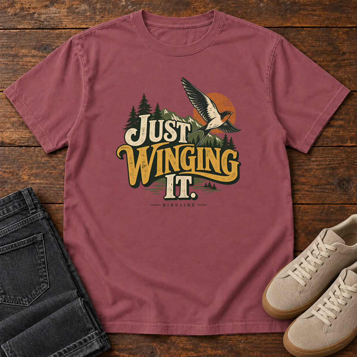 Brick Just Winging It T-Shirt with a flying bird, evergreens, an orange sun and cream and gold lettering; front flat lay on wood.