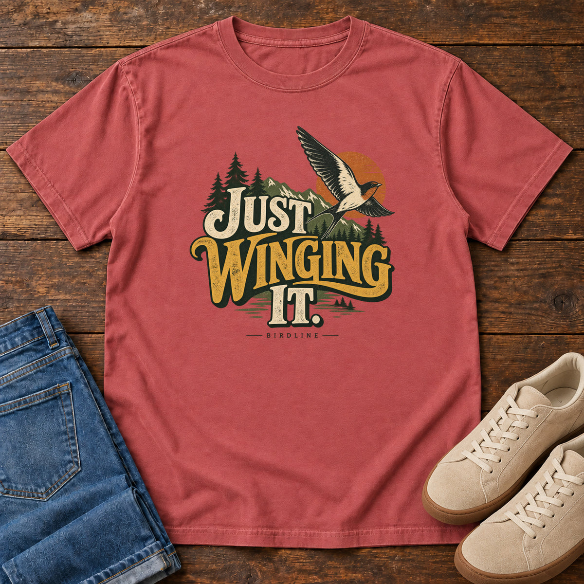 Crimson Just Winging It T-Shirt with a flying bird, evergreens, an orange sun and cream and gold lettering; front flat lay on wood.