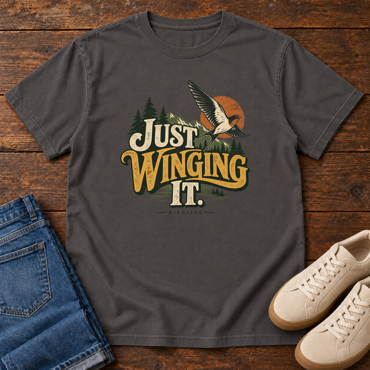 Graphite Just Winging It T-Shirt with a flying bird, evergreens, an orange sun and cream and gold lettering; front flat lay on wood.