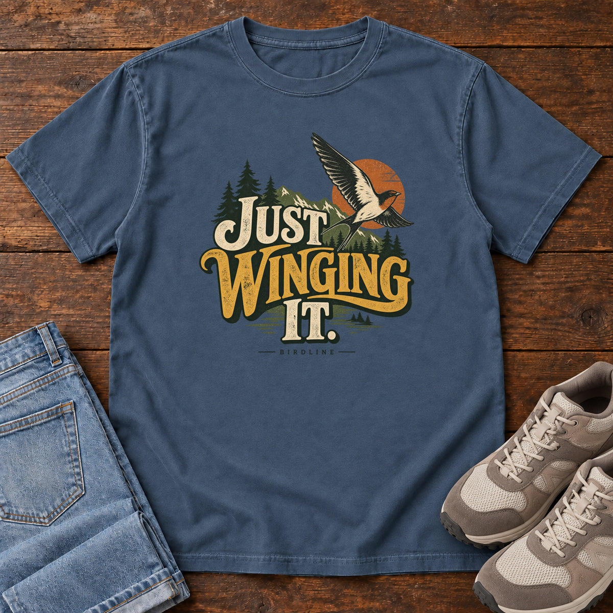 Midnight Just Winging It T-Shirt with a flying bird, evergreens, an orange sun and cream and gold lettering; front flat lay on wood.