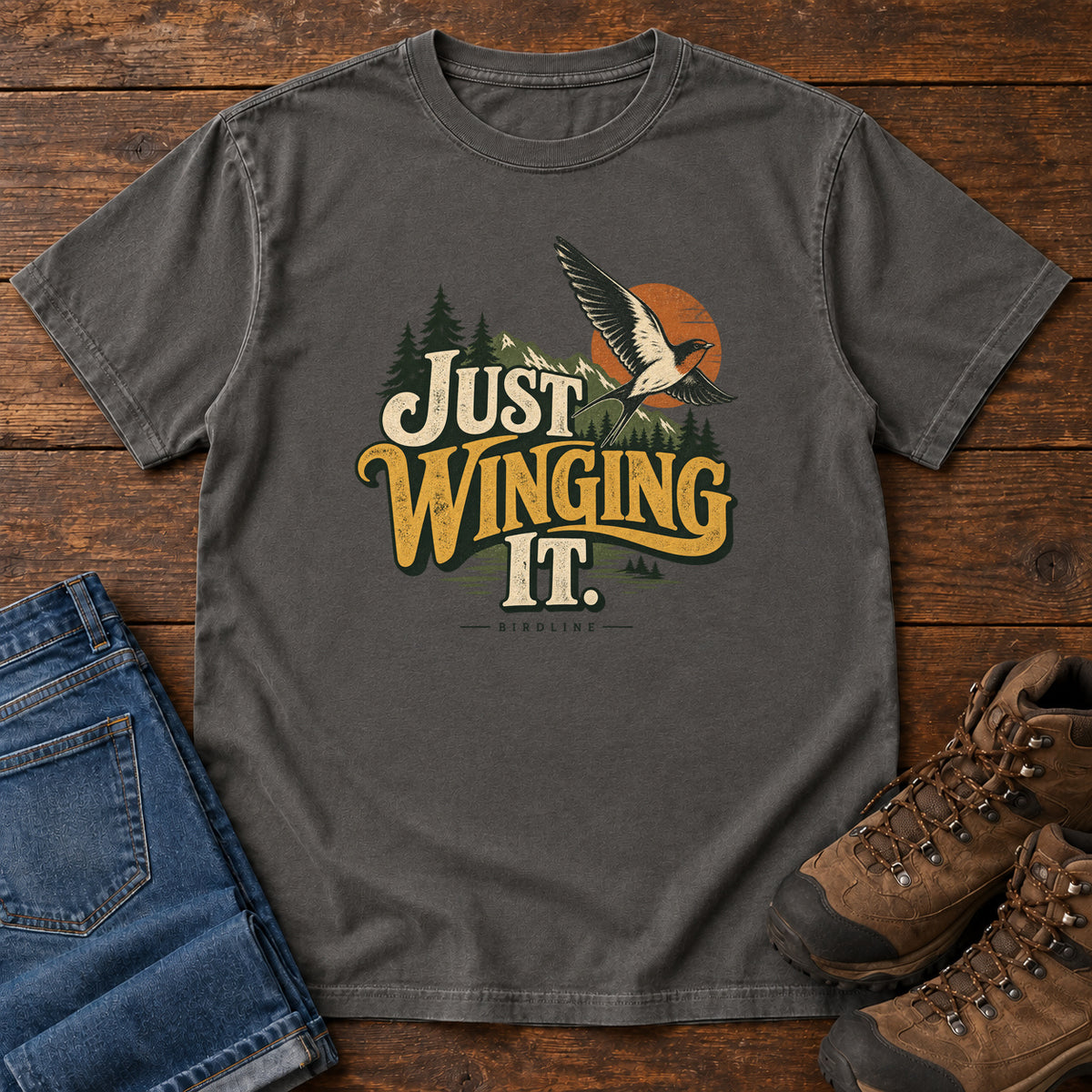 Pepper Just Winging It T-Shirt with a flying bird, evergreens, an orange sun and cream and gold lettering; front flat lay on wood.