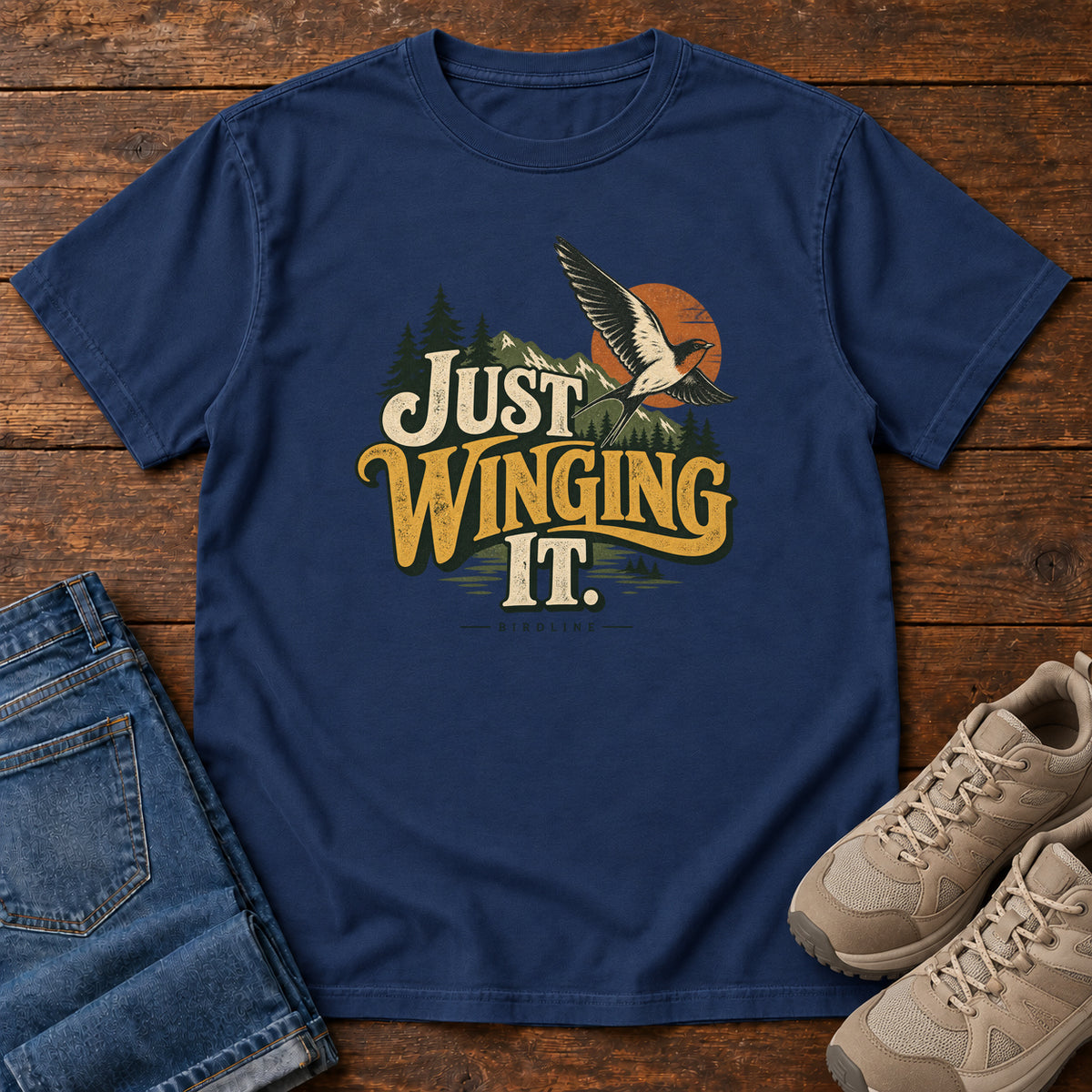 True Navy Just Winging It T-Shirt with a flying bird, evergreens, an orange sun and cream and gold lettering; front flat lay on wood.