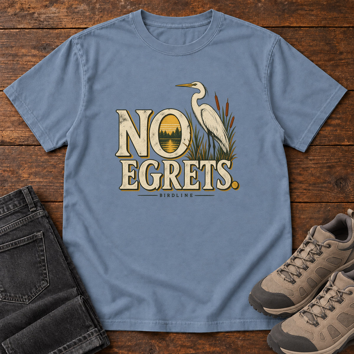 Blue Jean No Egrets. T-Shirt with a white long-necked wading bird beside reeds and lettering framing a sunset; front flat lay on wood.