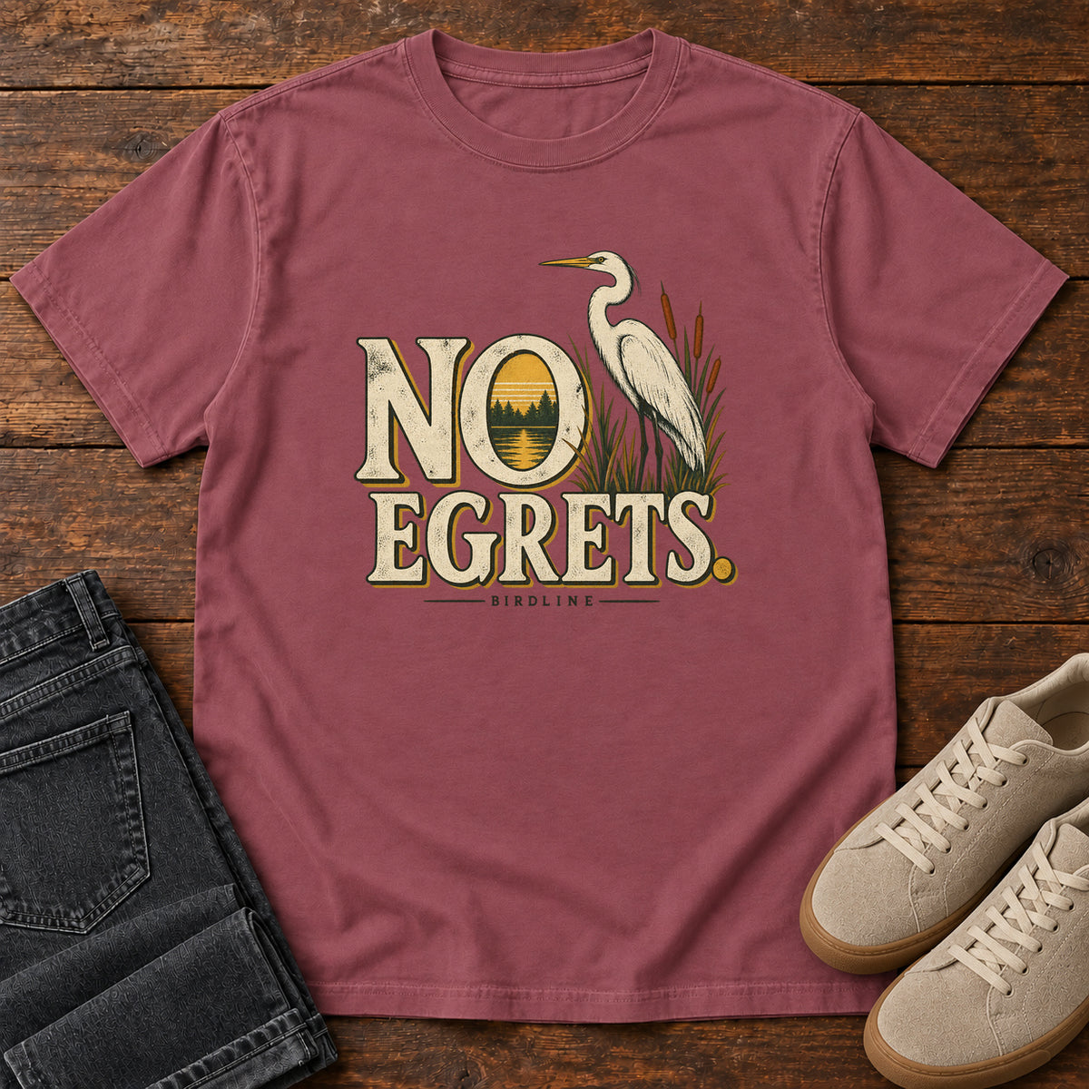 Brick No Egrets. T-Shirt with a white long-necked wading bird beside reeds and lettering framing a sunset; front flat lay on wood.
