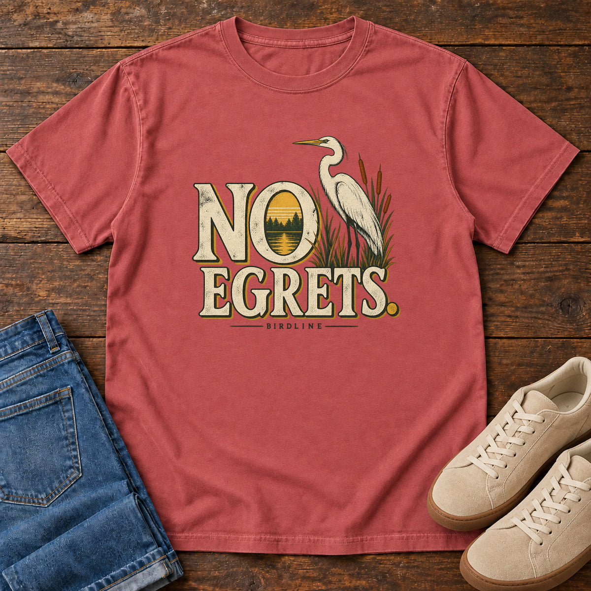 Crimson No Egrets. T-Shirt with a white long-necked wading bird beside reeds and lettering framing a sunset; front flat lay on wood.