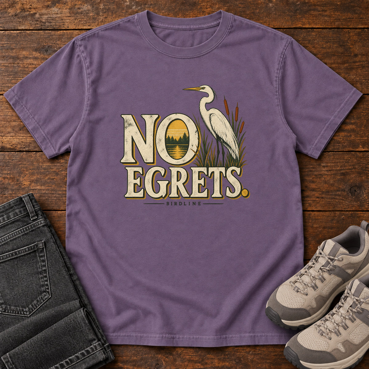 Grape No Egrets. T-Shirt with a white long-necked wading bird beside reeds and lettering framing a sunset; front flat lay on wood.