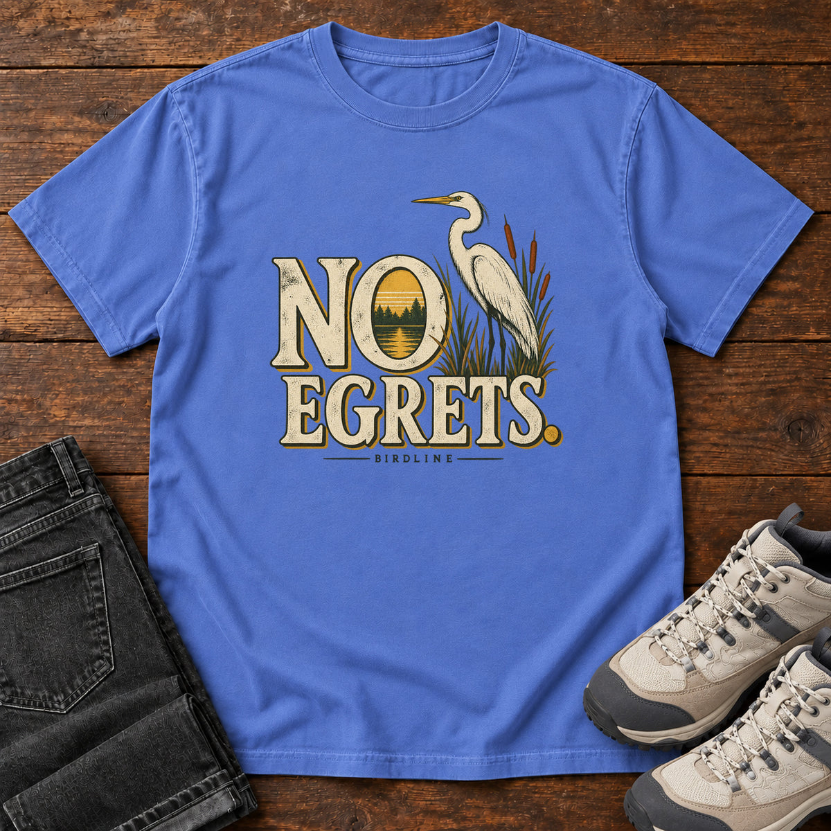 Mystic Blue No Egrets. T-Shirt with a white long-necked wading bird beside reeds and lettering framing a sunset; front flat lay on wood.
