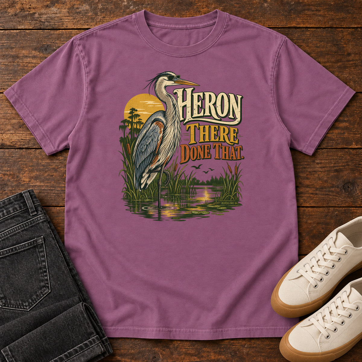 Berry Heron There Done That T-Shirt with a long-legged gray bird standing in marsh water beside sunset and cream and orange lettering, front flat lay on wood.