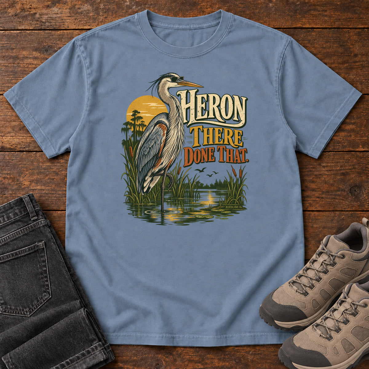 Blue Jean Heron There Done That T-Shirt with a long-legged gray bird standing in marsh water beside sunset and cream and orange lettering, front flat lay on wood.