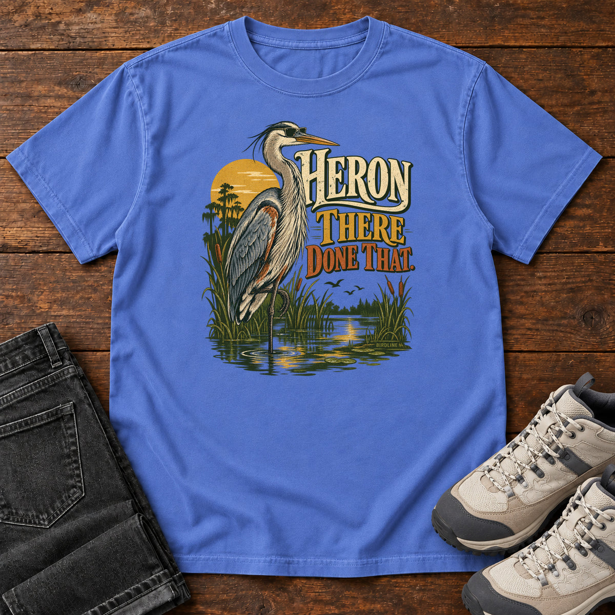 Mystic Blue Heron There Done That T-Shirt with a long-legged gray bird standing in marsh water beside sunset and cream and orange lettering, front flat lay on wood.