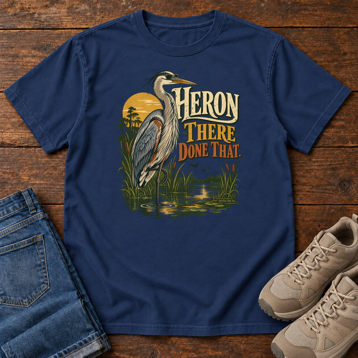 True Navy Heron There Done That T-Shirt with a long-legged gray bird standing in marsh water beside sunset and cream and orange lettering, front flat lay on wood.