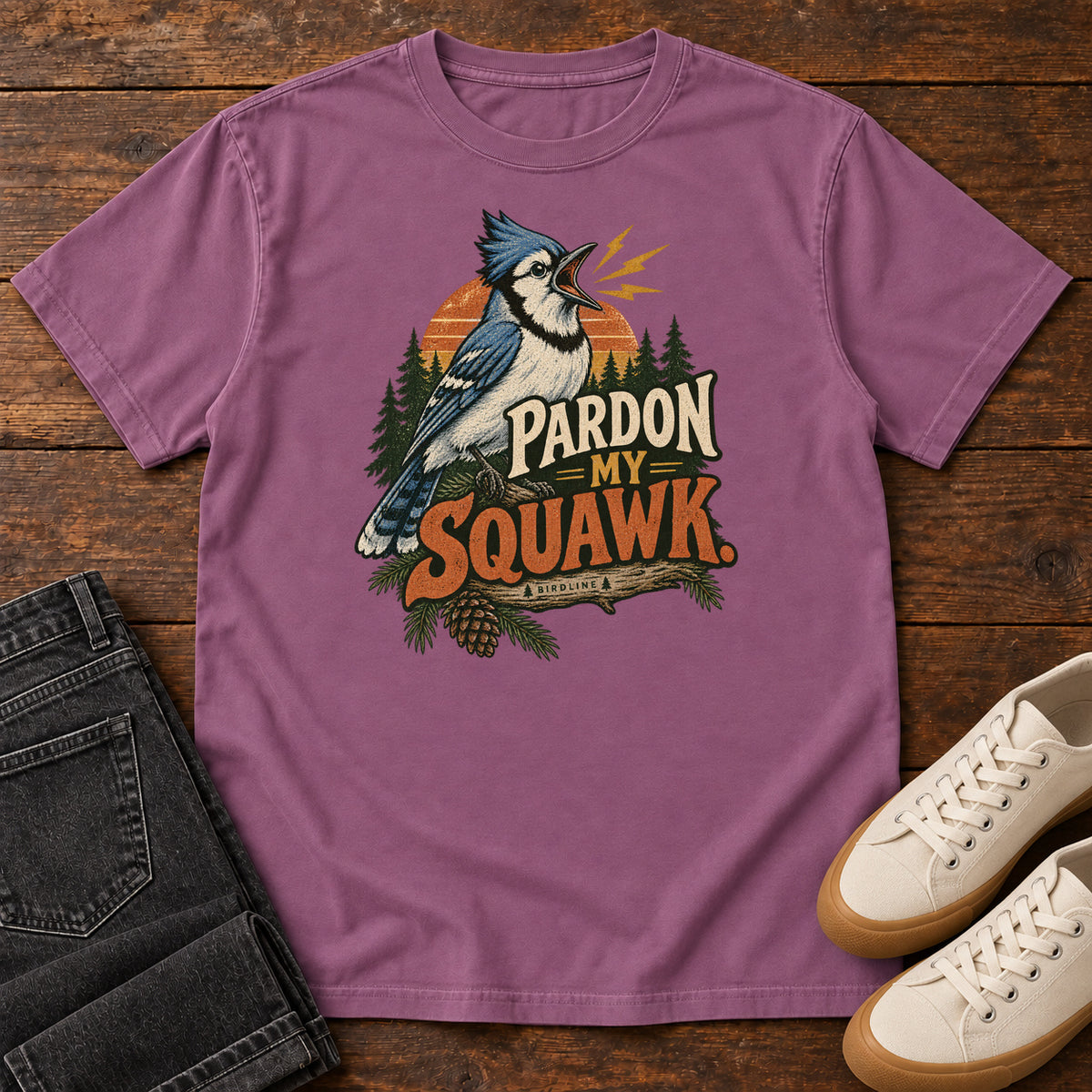 Pardon My Squawk T-Shirt in Berry, front laid flat on wood, featuring a blue-and-white crested bird with an open beak, orange sound marks, pine trees and bold lettering.