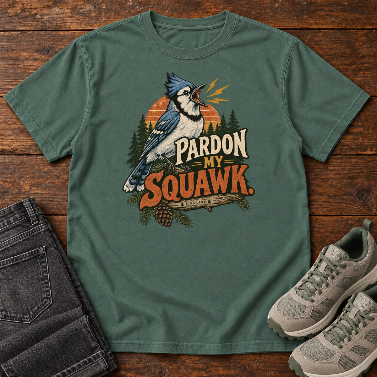 Pardon My Squawk T-Shirt in Blue Spruce, front laid flat on wood, featuring a blue-and-white crested bird with an open beak, orange sound marks, pine trees and bold lettering.