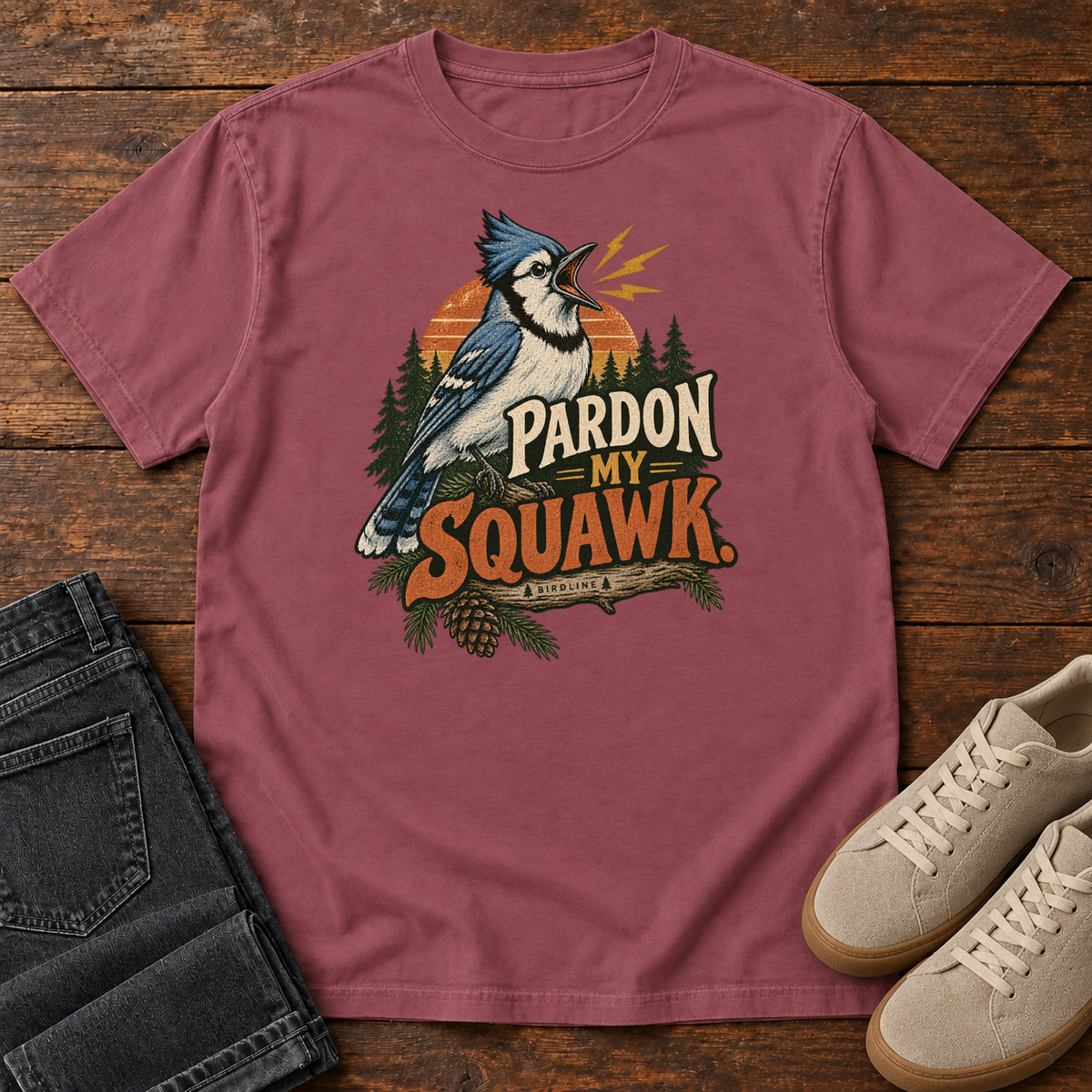 Pardon My Squawk T-Shirt in Brick, front laid flat on wood, featuring a blue-and-white crested bird with an open beak, orange sound marks, pine trees and bold lettering.