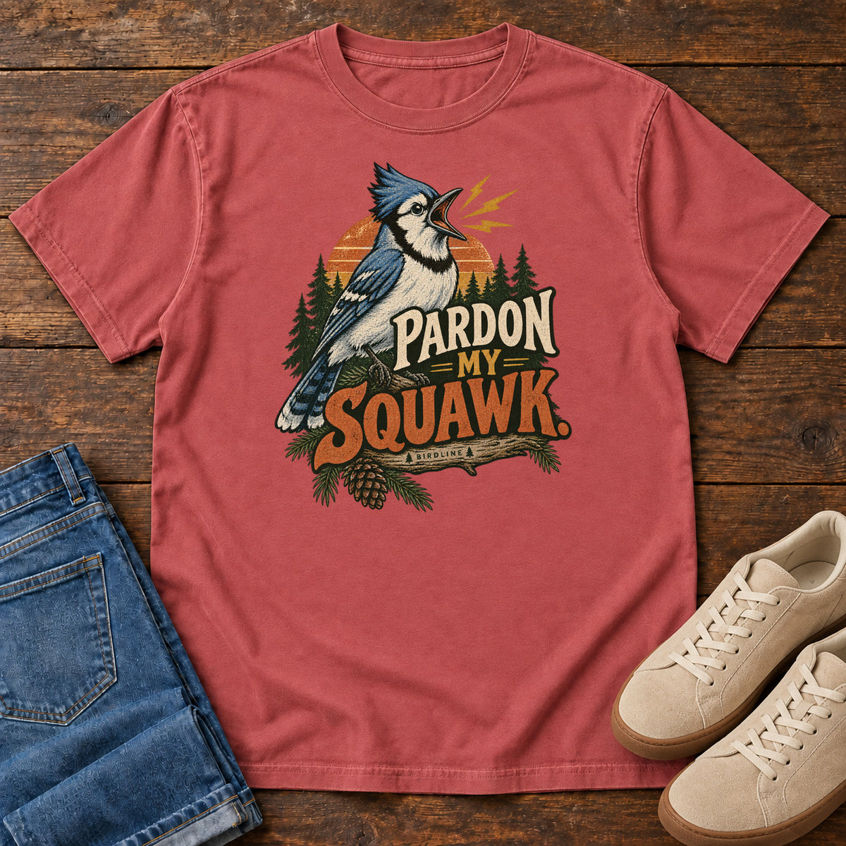 Pardon My Squawk T-Shirt in Crimson, front laid flat on wood, featuring a blue-and-white crested bird with an open beak, orange sound marks, pine trees and bold lettering.