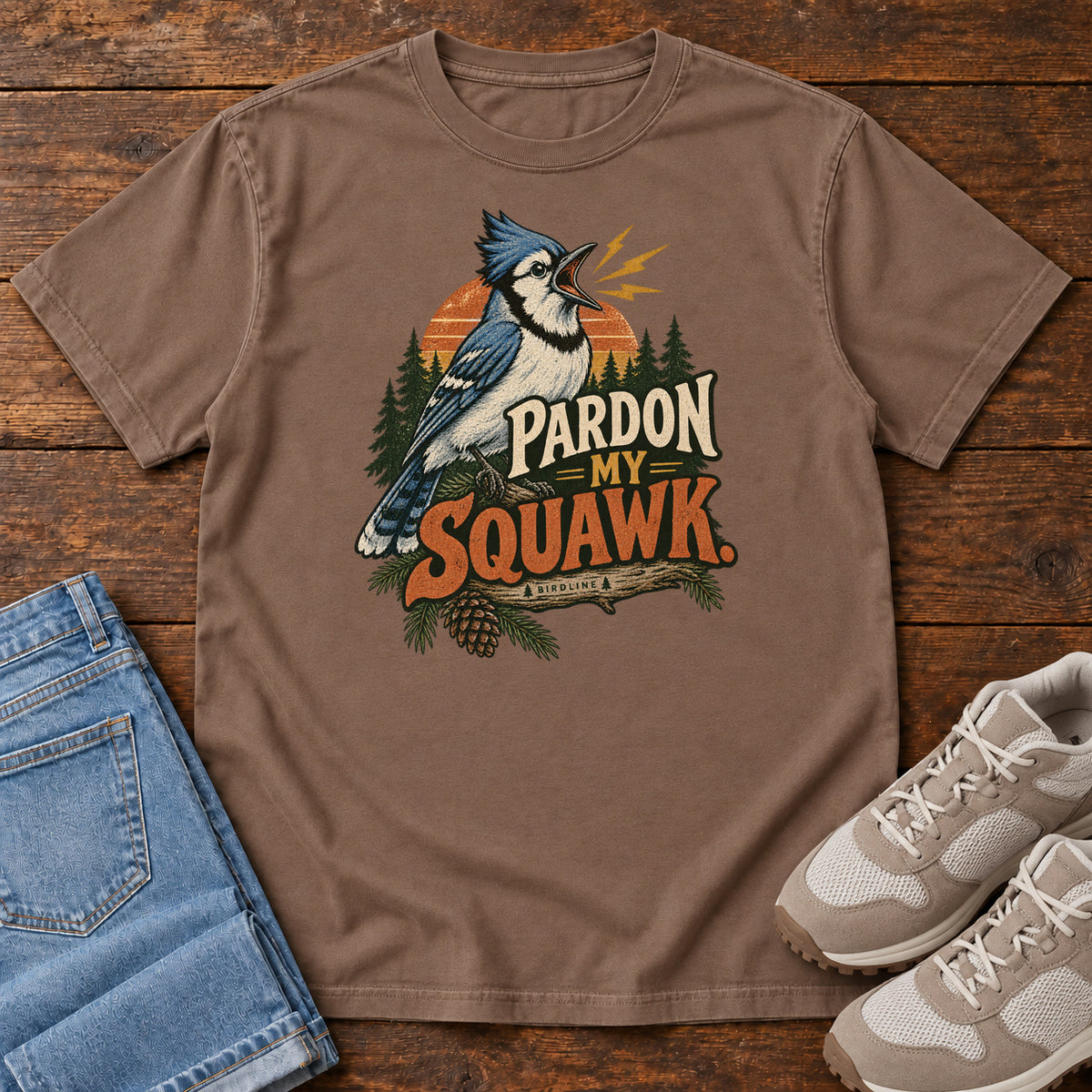 Pardon My Squawk T-Shirt in Espresso, front laid flat on wood, featuring a blue-and-white crested bird with an open beak, orange sound marks, pine trees and bold lettering.