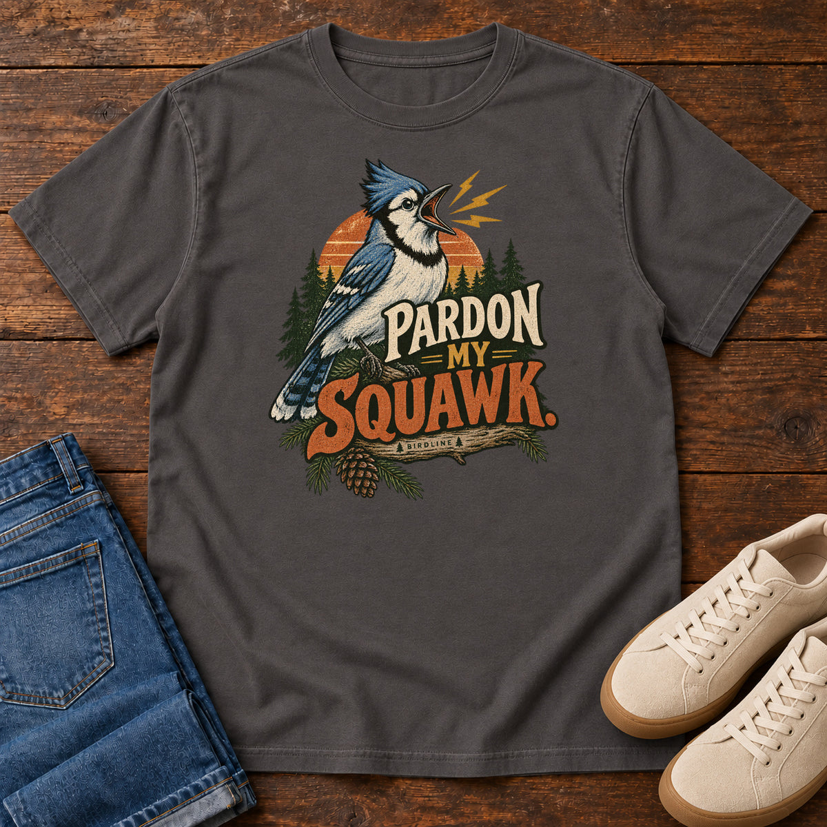 Pardon My Squawk T-Shirt in Graphite, front laid flat on wood, featuring a blue-and-white crested bird with an open beak, orange sound marks, pine trees and bold lettering.