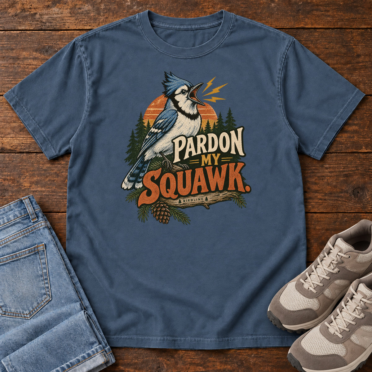 Pardon My Squawk T-Shirt in Midnight, front laid flat on wood, featuring a blue-and-white crested bird with an open beak, orange sound marks, pine trees and bold lettering.