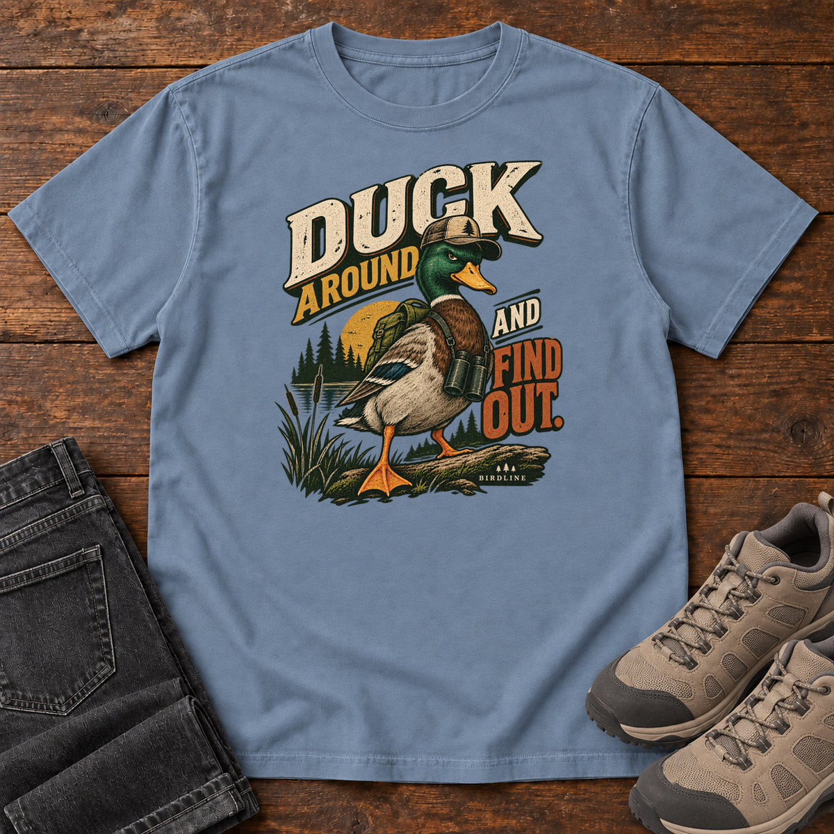 Blue Jean Duck Around and Find Out T-Shirt with a green-headed duck in a hat and vest beside marsh reeds and bold lettering, front flat lay on wood.