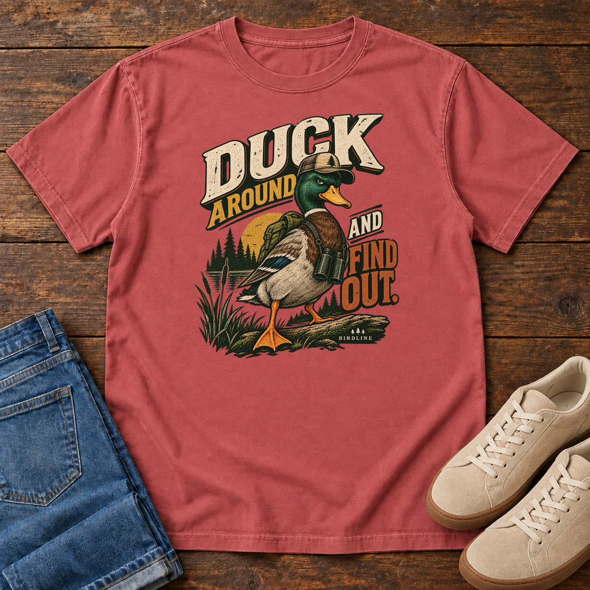 Crimson Duck Around and Find Out T-Shirt with a green-headed duck in a hat and vest beside marsh reeds and bold lettering, front flat lay on wood.
