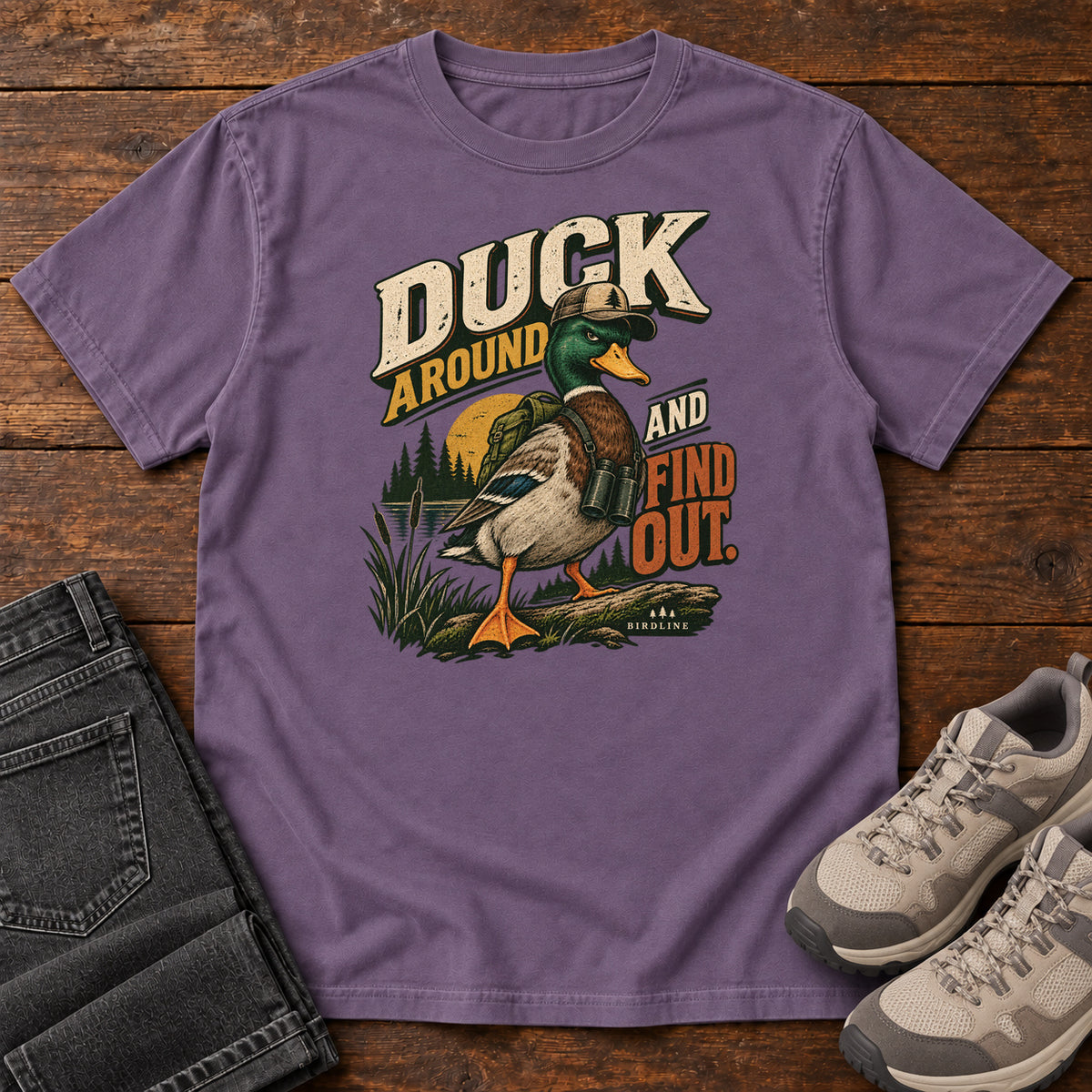 Grape Duck Around and Find Out T-Shirt with a green-headed duck in a hat and vest beside marsh reeds and bold lettering, front flat lay on wood.