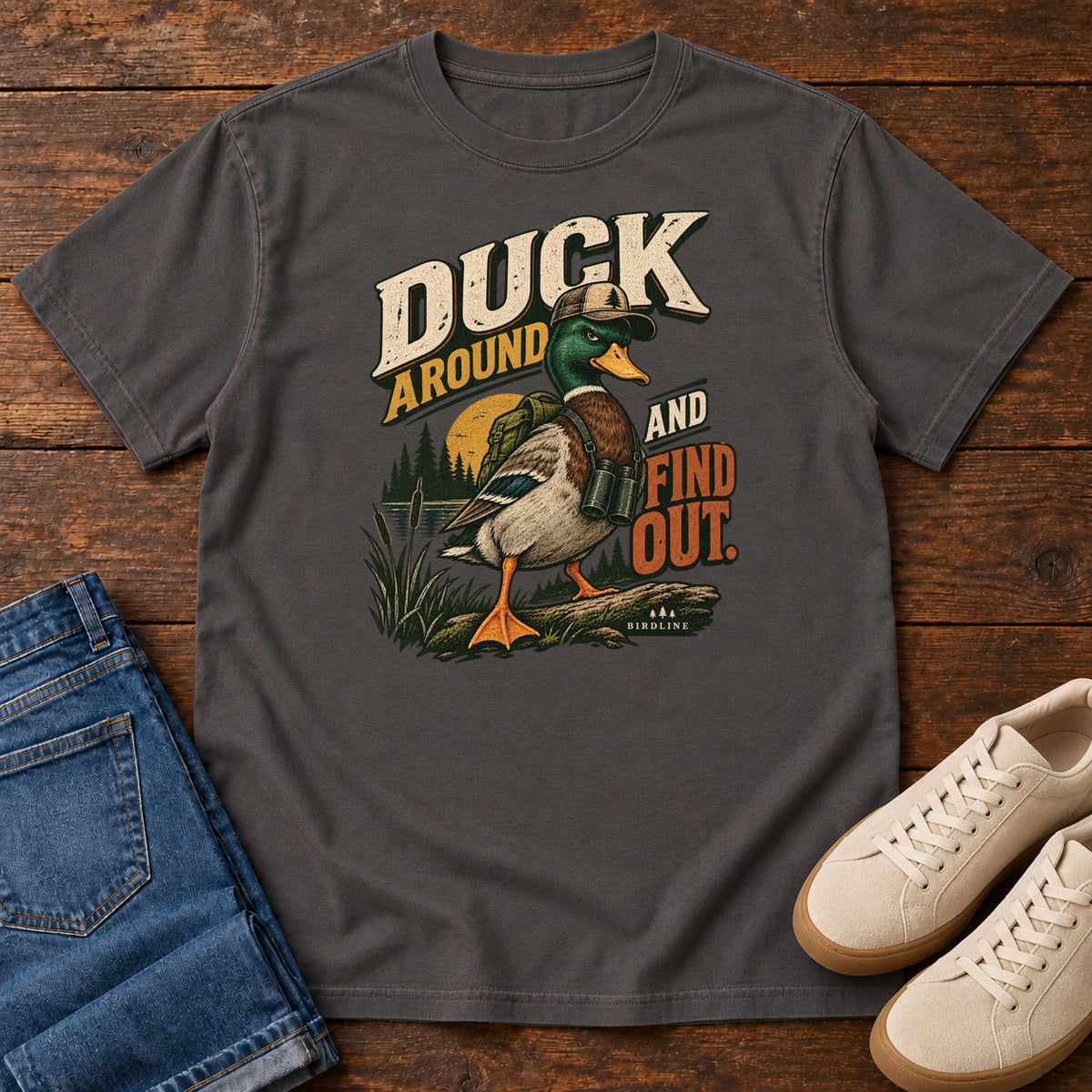 Graphite Duck Around and Find Out T-Shirt with a green-headed duck in a hat and vest beside marsh reeds and bold lettering, front flat lay on wood.