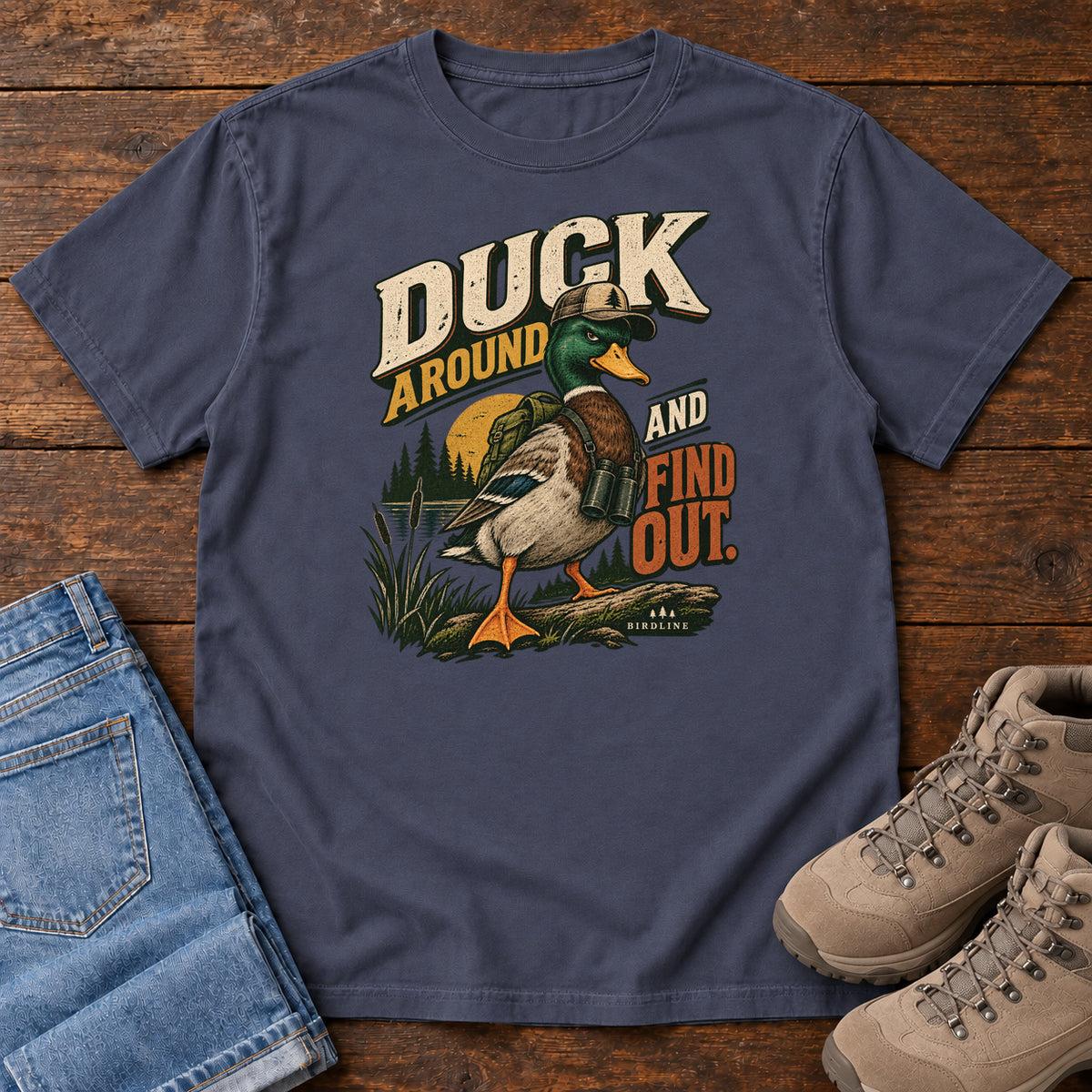 Navy Duck Around and Find Out T-Shirt with a green-headed duck in a hat and vest beside marsh reeds and bold lettering, front flat lay on wood.