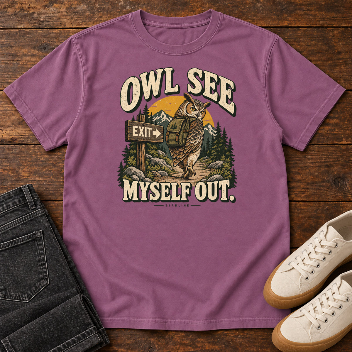 Berry Owl See Myself Out T-Shirt with an owl wearing a backpack beside an Exit sign on a woodland trail; front flat lay on wood.