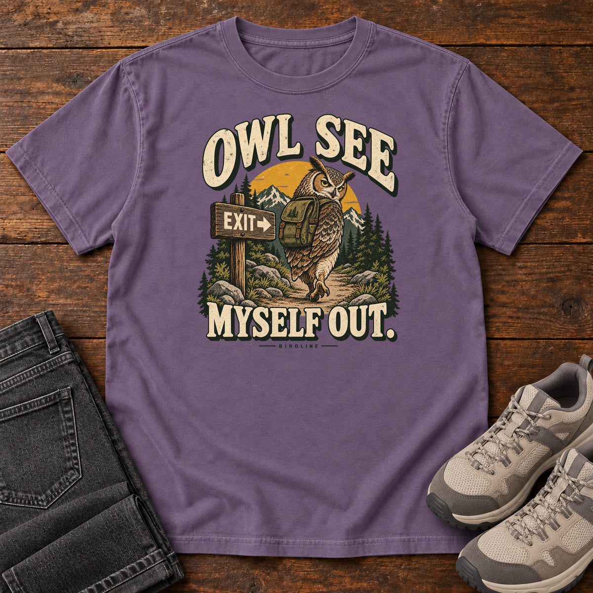Grape Owl See Myself Out T-Shirt with an owl wearing a backpack beside an Exit sign on a woodland trail; front flat lay on wood.