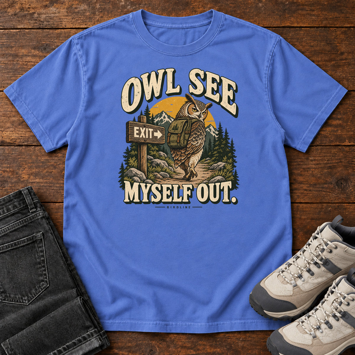 Mystic Blue Owl See Myself Out T-Shirt with an owl wearing a backpack beside an Exit sign on a woodland trail; front flat lay on wood.